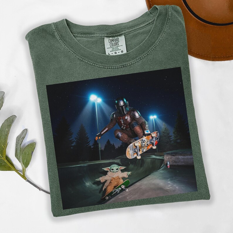 May include: Green Comfort Colors t-shirt featuring a graphic of The Mandalorian and Grogu skateboarding in a skate park at night. The image shows the characters mid-air and on skateboards. The shirt has a crew neck and short sleeves.