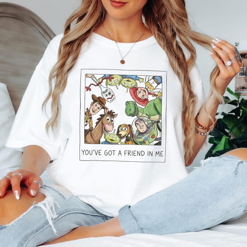 Pode incluir: Camiseta branca com uma ilustra&ccedil;&atilde;o colorida de personagens de Toy Story, incluindo Woody e Buzz Lightyear. O texto "YOU'VE GOT A FRIEND IN ME" &eacute; impresso abaixo da imagem, em um ret&acirc;ngulo.