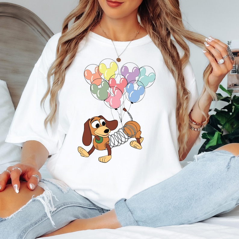 May include: White t-shirt featuring a cartoon illustration of Slinky Dog from Toy Story holding a bundle of Mickey Mouse-shaped balloons in various colors. The design is centered on the shirt.