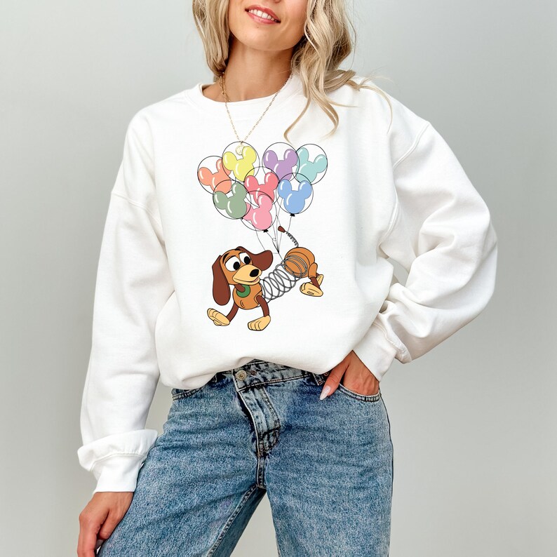 May include: White sweatshirt featuring a cartoon dachshund dog holding colorful Mickey Mouse-shaped balloons. The dog has a spring-like body and is in a playful pose. The balloons are in shades of yellow, orange, pink, green, purple, and blue.