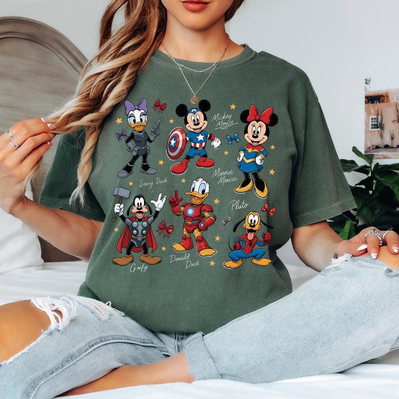 May include: Green t-shirt featuring cartoon characters Daisy Duck, Mickey Mouse, Minnie Mouse, Goofy, Donald Duck, and Pluto, each in superhero-themed costumes. The names of the characters are printed below them. Gold stars and red bows are scattered around the characters.