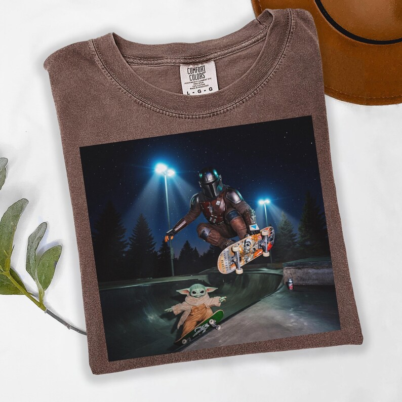 May include: Brown Comfort Colors t-shirt featuring a graphic of a character in Mandalorian armor skateboarding, with Baby Yoda on a skateboard in a skate park. The image is set at night with bright lights.