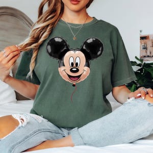 Puede incluir: Camiseta verde con un dise&ntilde;o de globo de Mickey Mouse. El globo tiene orejas negras, una cara sonriente y una cuerda roja. La camiseta es de color verde liso.