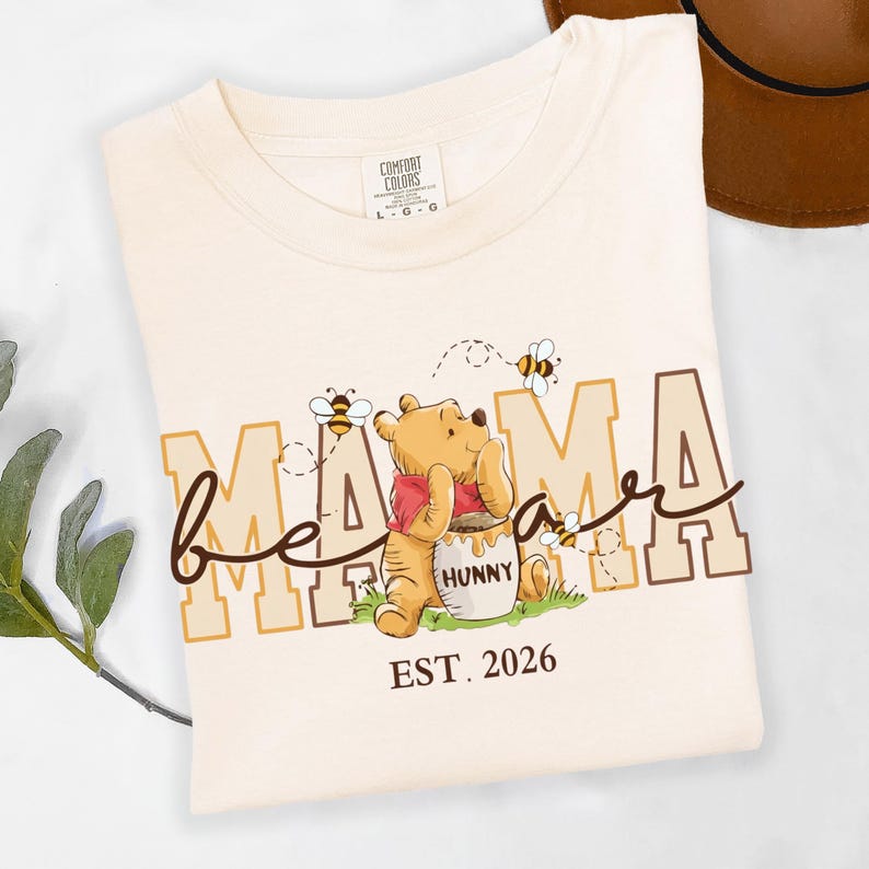 May include: Cream-colored t-shirt with a graphic featuring Winnie the Pooh, bees, and the words "Mama Bee" and "EST. 2026". The design includes a cartoon bear, honey pot, and bees. The shirt is folded, with a hat visible in the background.
