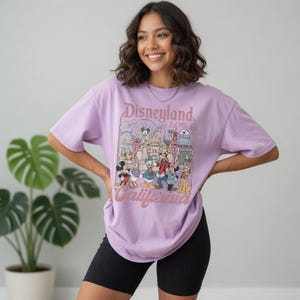 May include: A lilac t-shirt with a Disneyland graphic featuring Mickey Mouse, Donald Duck, and other characters. The graphic includes the text "Disneyland Est 1955 California". The shirt is worn with black shorts.