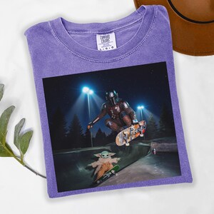 May include: Purple Comfort Colors t-shirt featuring a graphic of The Mandalorian and Grogu skateboarding in a skate park at night. The image shows the characters mid-air and on a skateboard. The shirt has a crew neck and short sleeves.