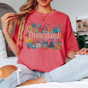 May include: A coral-colored t-shirt featuring a colorful Disneyland Resort graphic. The graphic includes the Disneyland logo and various park icons in shades of blue, red, yellow, and green. The shirt has a relaxed fit.