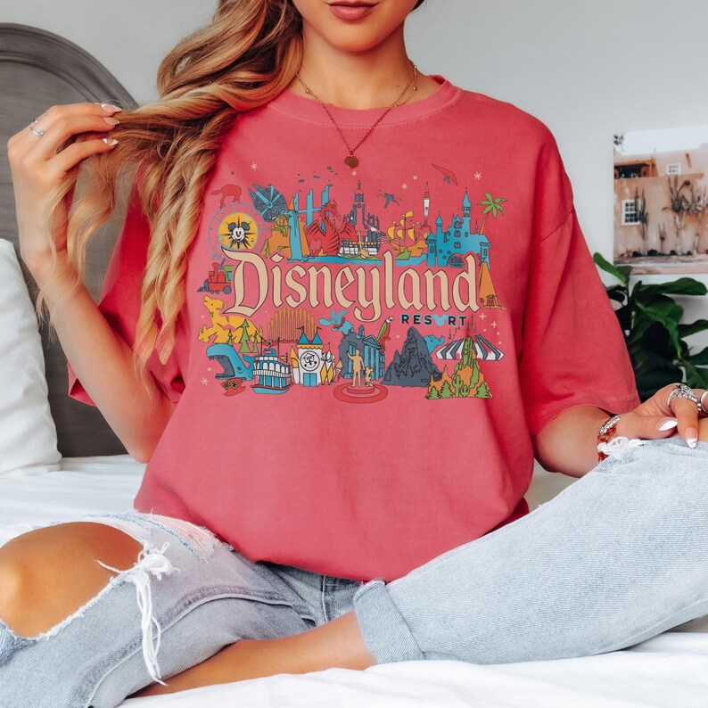 May include: A coral-colored t-shirt featuring a colorful Disneyland Resort graphic. The graphic includes the Disneyland logo and various park icons in shades of blue, red, yellow, and green. The shirt has a relaxed fit.