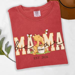 May include: Red t-shirt with the words "Bee Mama" in a whimsical font, featuring a cartoon bear and bees. The bear is holding a honey pot. The text "EST. 2026" is printed below the graphic. The shirt has a Comfort Colors tag.