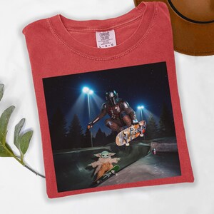 May include: Red t-shirt with a graphic featuring The Mandalorian and Grogu skateboarding in a skate park at night. The image includes the text "COMFORT COLORS" on the upper left of the shirt.
