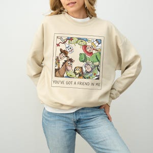 Pode incluir: Camisola bege com um gr&aacute;fico emoldurado de personagens de Toy Story. O gr&aacute;fico inclui Woody, Buzz Lightyear, Rex e outros personagens. O texto "YOU'VE GOT A FRIEND IN ME" est&aacute; impresso abaixo. A camisola &eacute; de cor neutra.