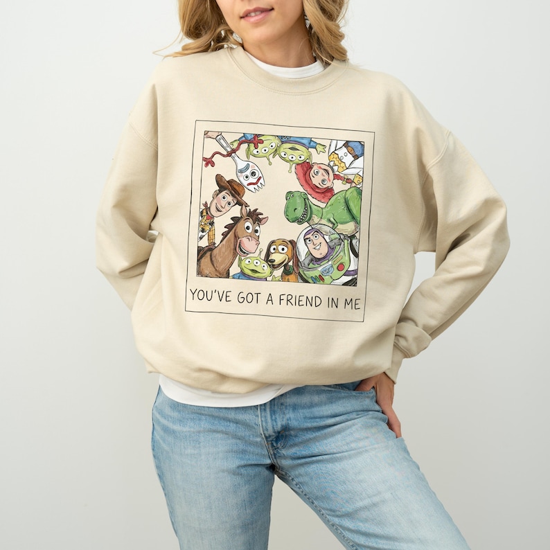 Pode incluir: Camisola bege com um gr&aacute;fico emoldurado de personagens de Toy Story. O gr&aacute;fico inclui Woody, Buzz Lightyear, Rex e outros personagens. O texto "YOU'VE GOT A FRIEND IN ME" est&aacute; impresso abaixo. A camisola &eacute; de cor neutra.