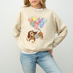 May include: Beige sweatshirt featuring a cartoon dachshund holding Mickey Mouse-shaped balloons in various colors. The dog has a spring-like body and is in a playful pose. The sweatshirt is a neutral color, and the design is centered on the front.