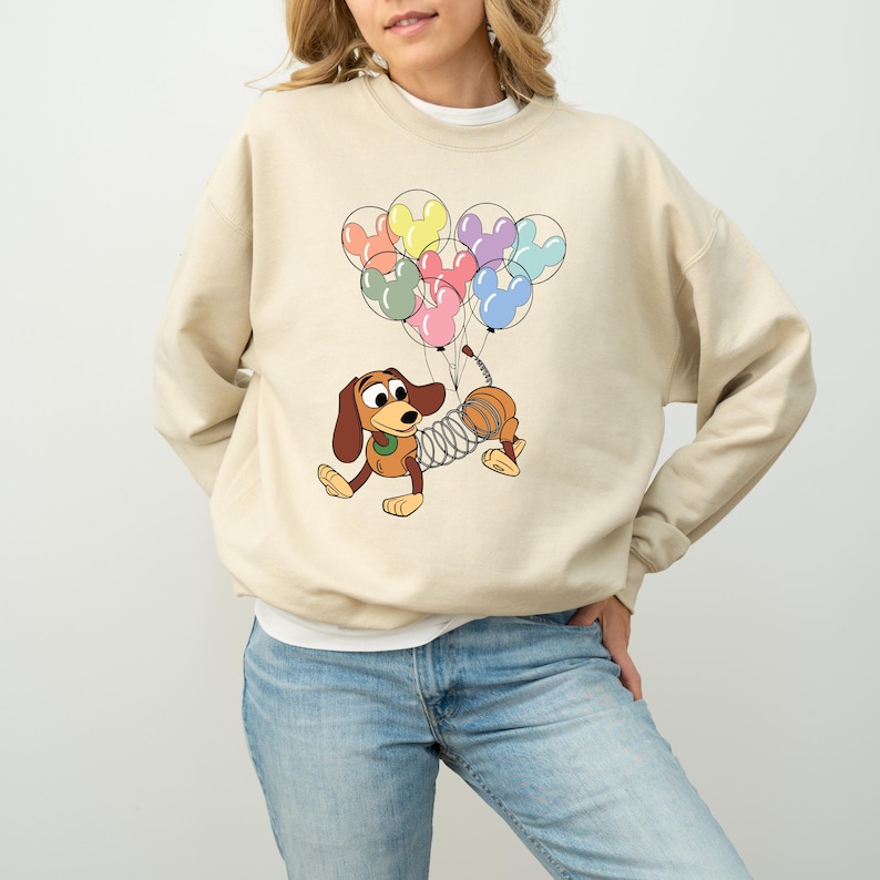 May include: Beige sweatshirt featuring a cartoon dachshund holding Mickey Mouse-shaped balloons in various colors. The dog has a spring-like body and is in a playful pose. The sweatshirt is a neutral color, and the design is centered on the front.