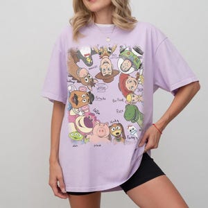 Colorful Toy Story characters with names in a playful retro style on a lavender t-shirt.