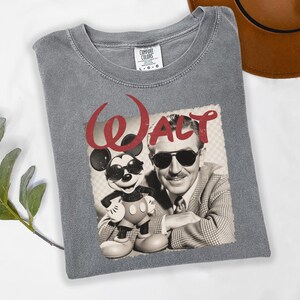 Puede incluir: Camiseta gris Comfort Colors con un gr&aacute;fico de Walt Disney y Mickey Mouse con gafas de sol. La palabra "Walt" est&aacute; impresa en rojo sobre la imagen. La camiseta est&aacute; doblada sobre una superficie blanca.