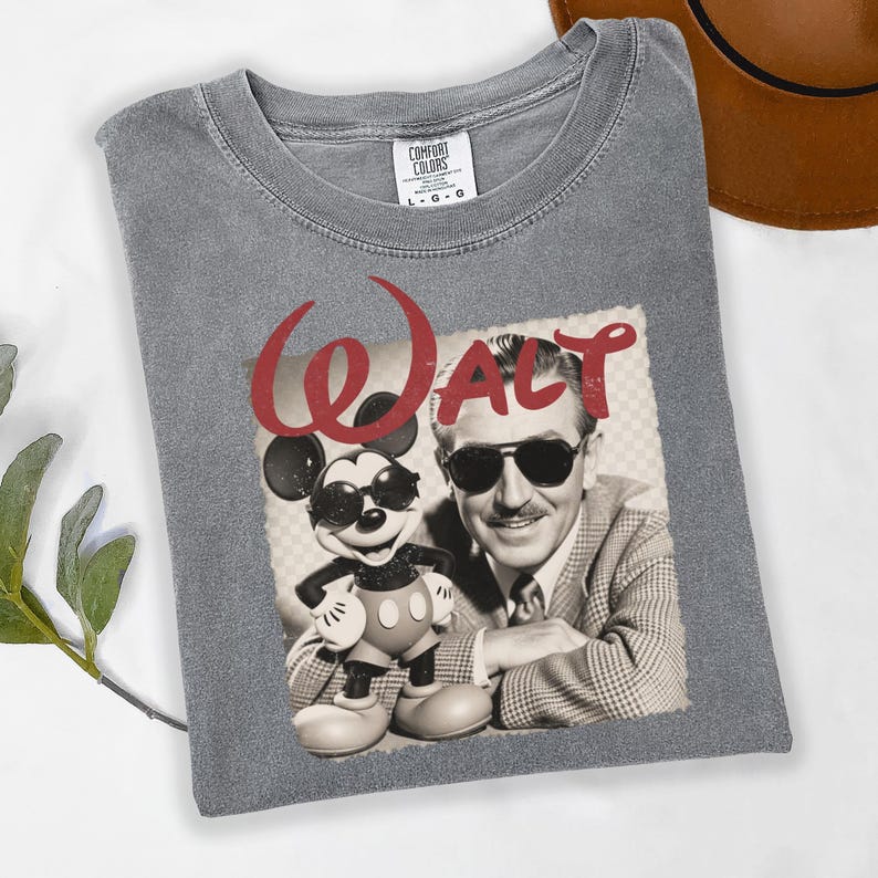 Puede incluir: Camiseta gris Comfort Colors con un gr&aacute;fico de Walt Disney y Mickey Mouse con gafas de sol. La palabra "Walt" est&aacute; impresa en rojo sobre la imagen. La camiseta est&aacute; doblada sobre una superficie blanca.