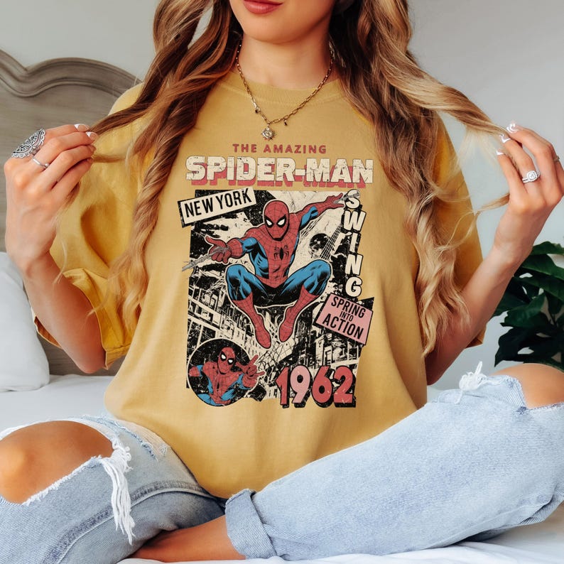 May include: Mustard yellow t-shirt featuring a vintage comic book graphic of Spider-Man. The design includes the text "THE AMAZING SPIDER-MAN NEW YORK SWING SPRING INTO ACTION 1962". The superhero is depicted in his classic red and blue suit.