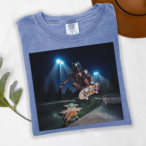 May include: Blue Comfort Colors t-shirt featuring a graphic of The Mandalorian and Grogu skateboarding in a skate park at night. The image includes the Comfort Colors logo.