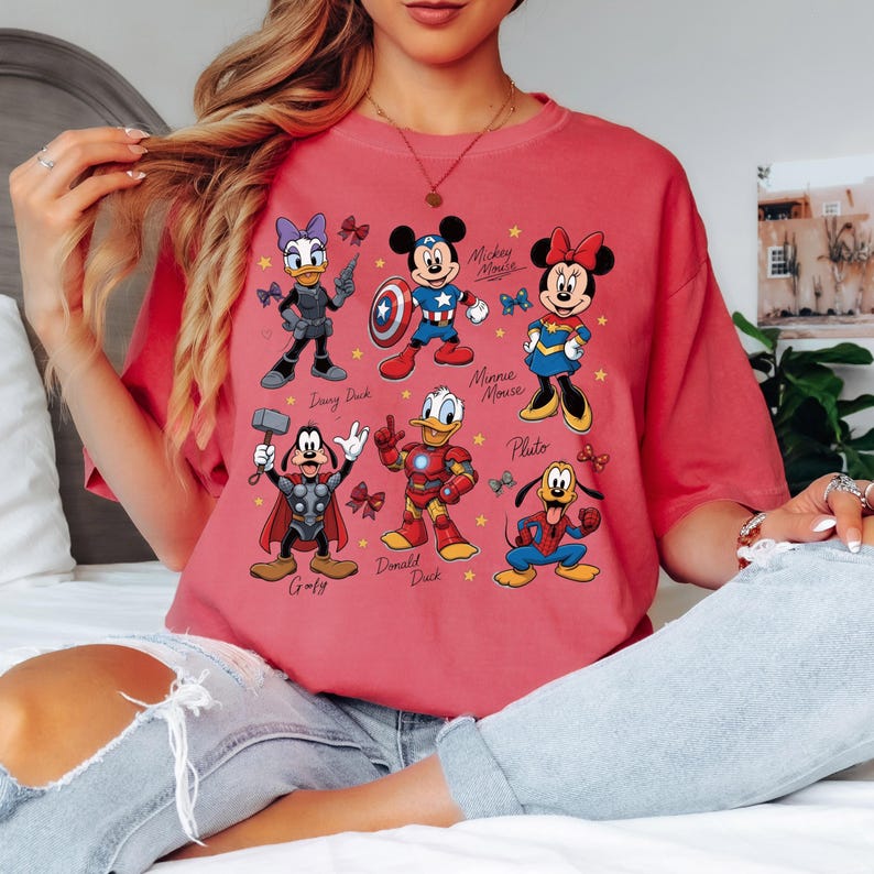 May include: A red t-shirt featuring cartoon characters Daisy Duck, Mickey Mouse, Minnie Mouse, Goofy, Donald Duck, and Pluto, each dressed as superheroes. The names of the characters are printed below them. The shirt is a comfortable, casual style.