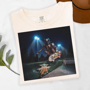 May include: Cream-colored t-shirt featuring a graphic of The Mandalorian and Grogu skateboarding in a skate park at night. The image is centered on the shirt, with the Comfort Colors logo visible at the top.