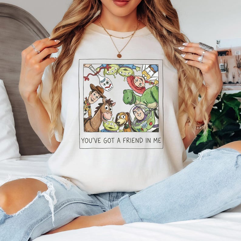 Pode incluir: Camiseta creme com uma ilustra&ccedil;&atilde;o emoldurada de personagens do filme de anima&ccedil;&atilde;o Toy Story. A imagem inclui Woody, Buzz Lightyear e outros personagens. O texto "YOU'VE GOT A FRIEND IN ME" est&aacute; impresso abaixo da ilustra&ccedil;&atilde;o.