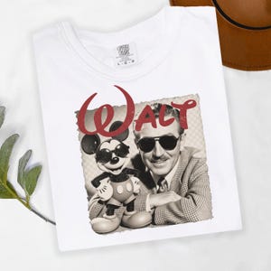 Puede incluir: Camiseta blanca con un gr&aacute;fico vintage de Walt Disney y Mickey Mouse con gafas de sol. La palabra "Walt" est&aacute; impresa en rojo encima de la imagen. La camiseta tiene cuello redondo y una etiqueta Comfort Colors.