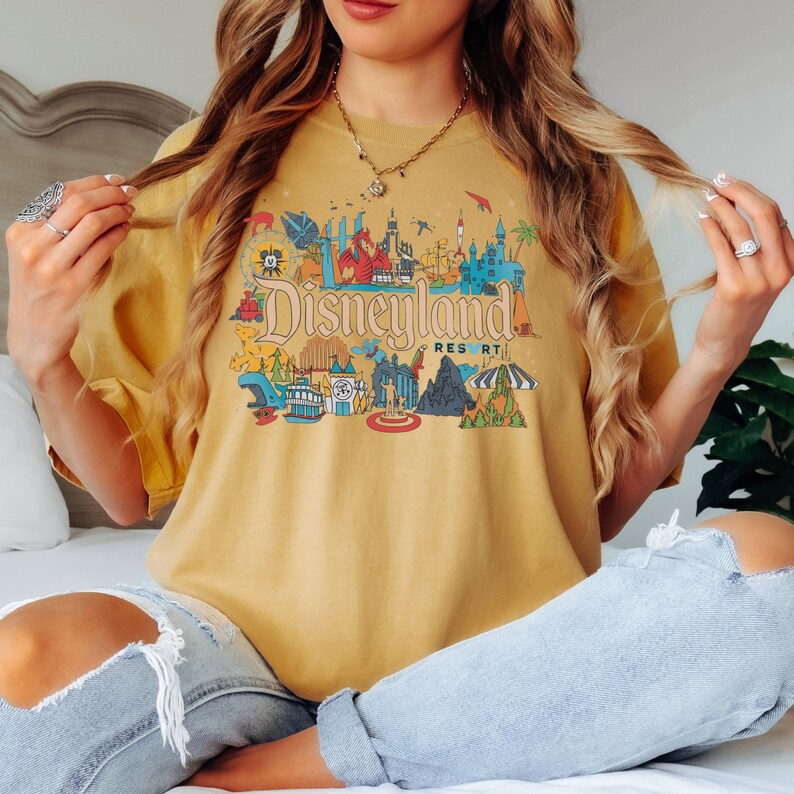 May include: Mustard yellow t-shirt featuring a colorful Disneyland Resort graphic. The graphic includes illustrations of iconic park landmarks, characters, and the Disneyland Resort logo. The shirt has short sleeves and a relaxed fit.