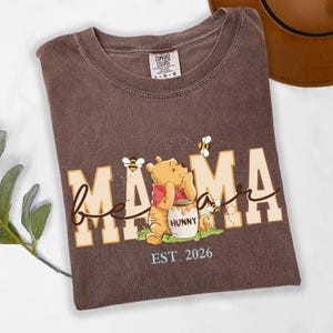 May include: Brown t-shirt with the words "bee mama" in a playful font, featuring a cartoon bear holding a honey pot. The shirt also includes the text "EST. 2026" and images of bees.