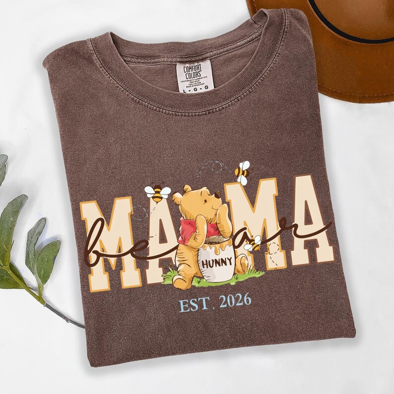 May include: Brown t-shirt with the words "bee mama" in a playful font, featuring a cartoon bear holding a honey pot. The shirt also includes the text "EST. 2026" and images of bees.
