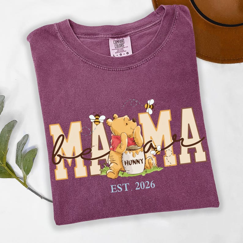 May include: A purple t-shirt with the words "bee mama" in a playful font, featuring a cartoon bear and bees. The bear is holding a honey pot. The text "EST. 2026" is below the graphic. The shirt is a Comfort Colors brand.