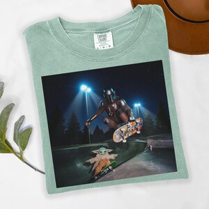 May include: Sage green t-shirt with a graphic featuring The Mandalorian and Grogu skateboarding in a skate park at night. The shirt has a crew neck and the Comfort Colors brand tag.