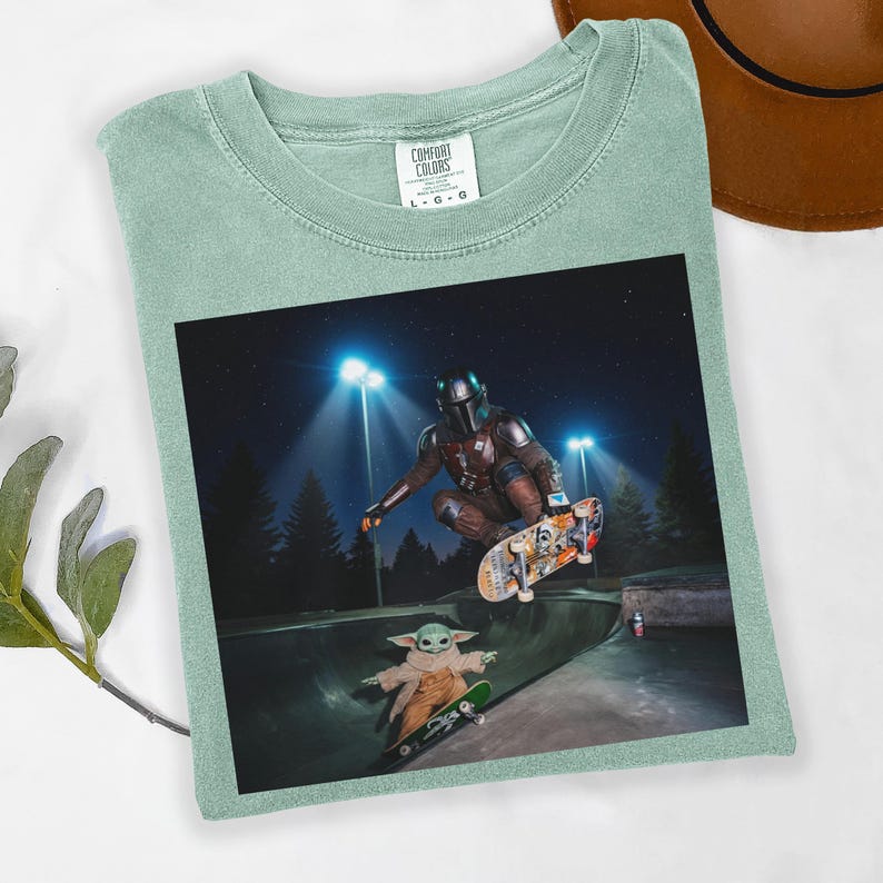 May include: Sage green t-shirt with a graphic featuring The Mandalorian and Grogu skateboarding in a skate park at night. The shirt has a crew neck and the Comfort Colors brand tag.