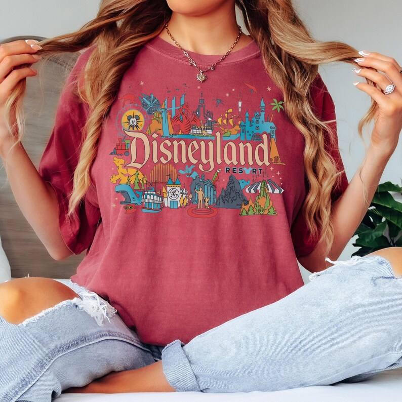 May include: A maroon t-shirt featuring a colorful Disneyland Resort graphic. The design includes various park icons and the Disneyland logo in a vintage style. The shirt is a comfortable fit and suitable for casual wear.