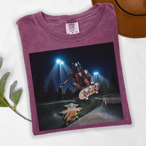 May include: A maroon Comfort Colors t-shirt featuring a graphic of The Mandalorian and Grogu skateboarding in a skate park at night. The image shows the characters mid-air and on a ramp. The shirt has a crew neck.