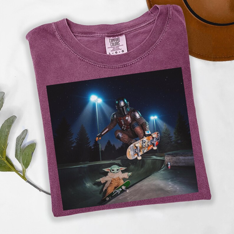 May include: A maroon Comfort Colors t-shirt featuring a graphic of The Mandalorian and Grogu skateboarding in a skate park at night. The image shows the characters mid-air and on a ramp. The shirt has a crew neck.