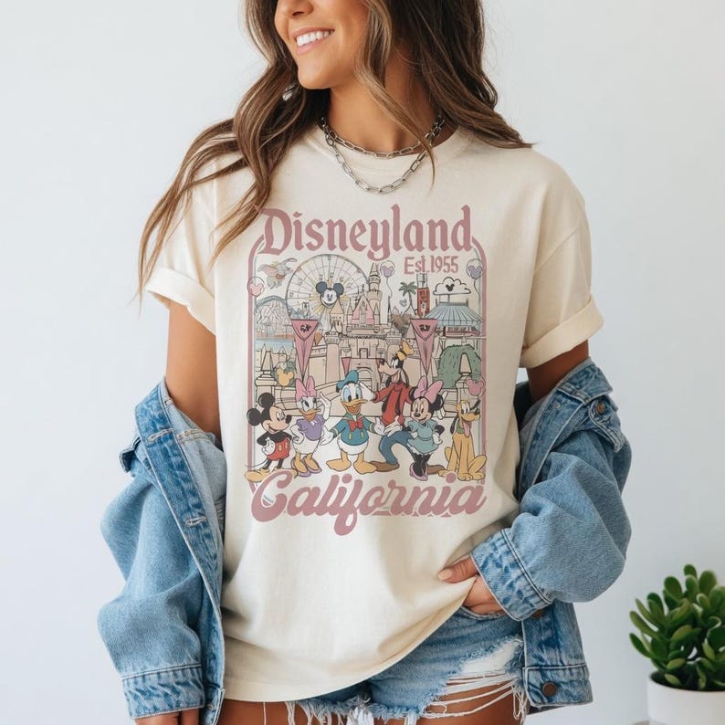 May include: Cream-coloured t-shirt with a Disneyland graphic featuring Mickey Mouse, Donald Duck, and other cartoon characters. The graphic includes the text "Disneyland Est. 1955 California" in a vintage style. The shirt is worn with a denim jacket.