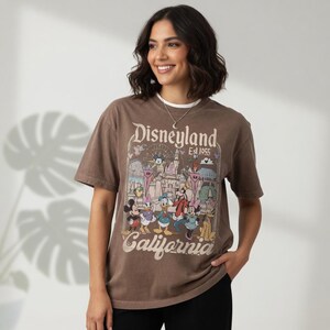 May include: A brown t-shirt with a Disneyland graphic featuring Mickey Mouse, Donald Duck, and other Disney characters. The shirt has the words "Disneyland Est. 1955 California" printed on it. The shirt is short-sleeved.