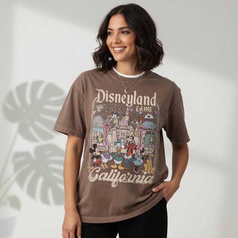 May include: A brown t-shirt with a Disneyland graphic featuring Mickey Mouse, Donald Duck, and other Disney characters. The shirt has the words "Disneyland Est. 1955 California" printed on it. The shirt is short-sleeved.