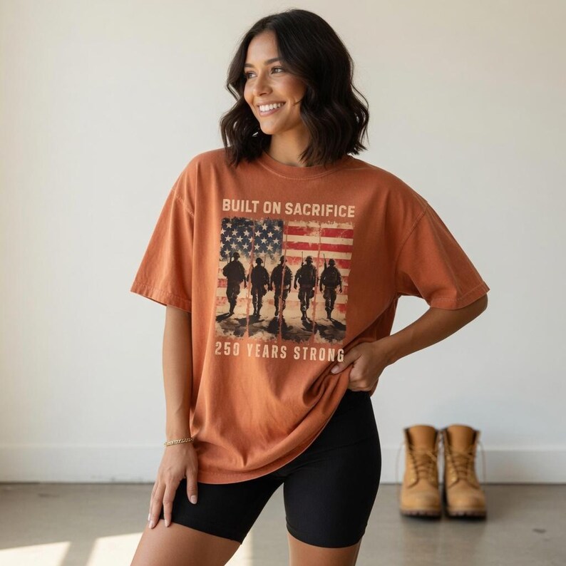 May include: Burnt orange t-shirt with a graphic of the American flag and silhouettes of soldiers. The text reads "BUILT ON SACRIFICE" and "250 YEARS STRONG". The shirt is paired with black shorts and tan boots.