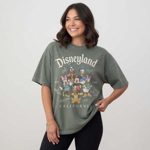 May include: An olive green t-shirt with the word "Disneyland" in a vintage font, and cartoon characters such as Mickey Mouse, Minnie Mouse, and Donald Duck. The shirt also includes the text "EST. 1955 CALIFORNIA".