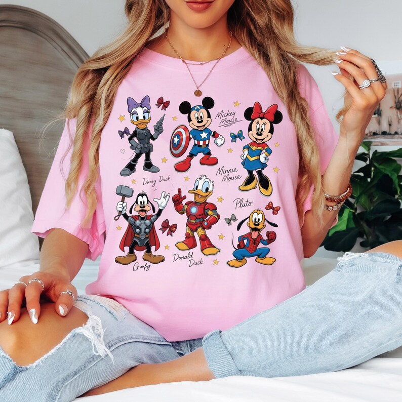 May include: A light pink t-shirt featuring cartoon characters Mickey Mouse, Minnie Mouse, Donald Duck, Daisy Duck, Goofy, and Pluto, each dressed as superheroes. The characters are surrounded by stars and bows.