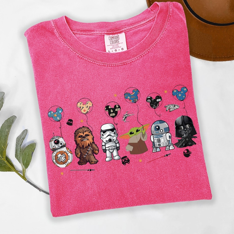 Puede incluir: Una camiseta rosa Comfort Colors con personajes de Star Wars: BB-8, Chewbacca, un Stormtrooper, Grogu, R2-D2 y Darth Vader. Sostienen globos con orejas de Mickey Mouse. La camiseta tiene cuello redondo.