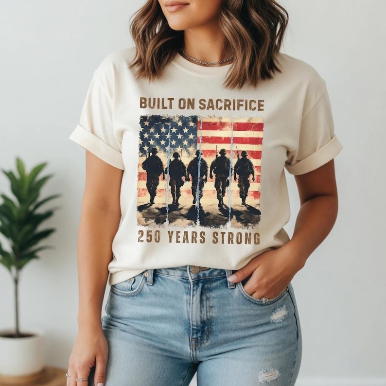 May include: Cream-colored t-shirt featuring a graphic of the American flag with silhouettes of soldiers. The text "BUILT ON SACRIFICE" and "250 YEARS STRONG" are printed above and below the flag design. The shirt has short sleeves.