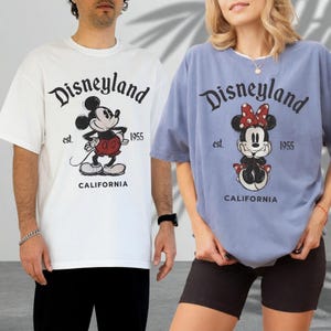 May include: Two short-sleeved t-shirts, one white and one periwinkle, featuring the Disneyland logo and a cartoon Mickey and Minnie Mouse. The shirts also include the text "est. 1955" and "CALIFORNIA".
