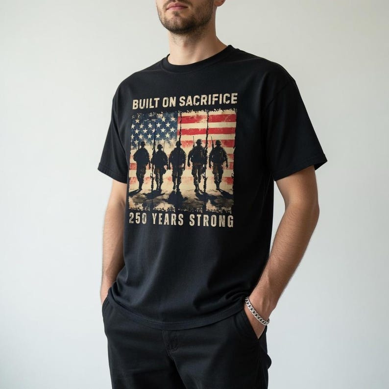 May include: Black t-shirt featuring an American flag design with soldiers and the text "BUILT ON SACRIFICE" and "250 YEARS STRONG." The graphic is in shades of red, white, and blue. The shirt is a classic crew neck style.