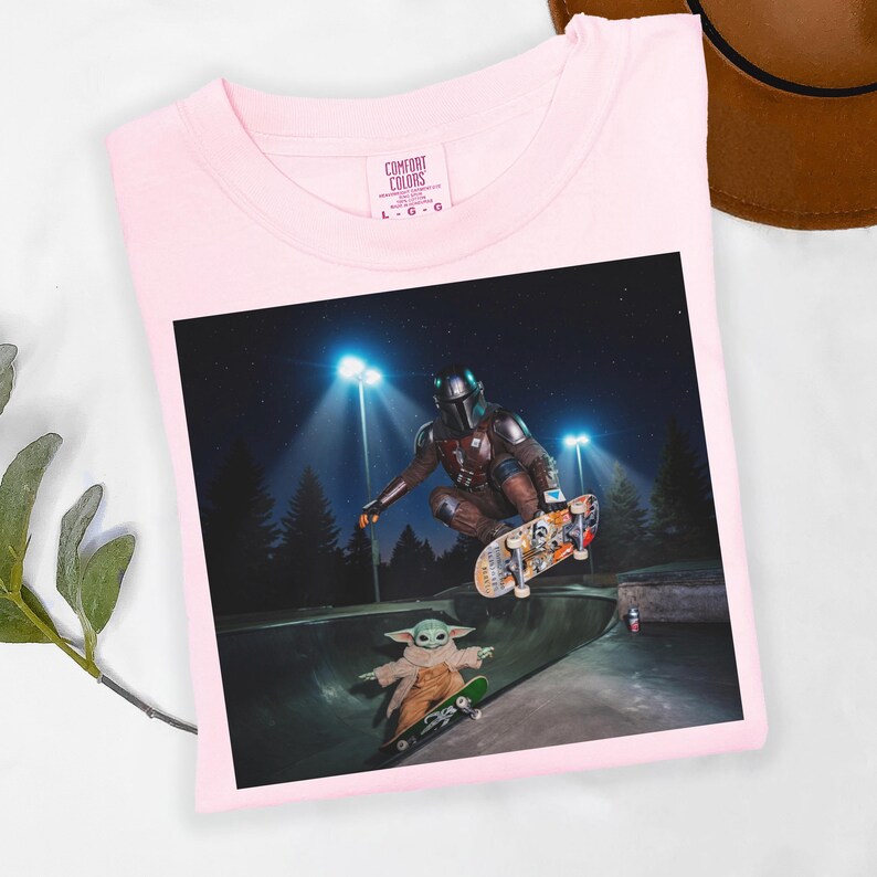 May include: Light pink Comfort Colors t-shirt featuring a graphic of The Mandalorian and Grogu skateboarding in a skate park at night. The Mandalorian is mid-air on a skateboard, while Grogu rides a smaller board below.