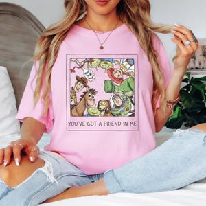 Pode incluir: T-shirt rosa claro com um gr&aacute;fico emoldurado de personagens de Toy Story, incluindo Woody, Buzz Lightyear e os alien&iacute;genas. O texto "YOU'VE GOT A FRIEND IN ME" est&aacute; impresso abaixo. A camisa &eacute; feita de material macio.