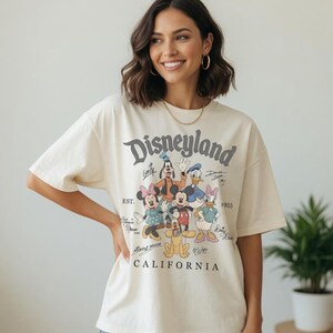 May include: Cream-coloured t-shirt with the word "Disneyland" in a vintage font, and cartoon characters such as Mickey Mouse, Minnie Mouse, and Donald Duck. The shirt also includes the text "California" and "EST. 1955".