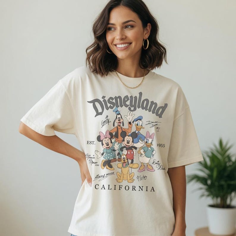 May include: Cream-coloured t-shirt with the word "Disneyland" in a vintage font, and cartoon characters such as Mickey Mouse, Minnie Mouse, and Donald Duck. The shirt also includes the text "California" and "EST. 1955".